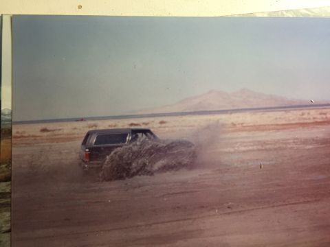 1988 Ford Bronco - A dark-colored Ford Bronco is kicking up a large amount of dirt and dust as it turns sharply on a dry, sandy plain. In the background, dist…
