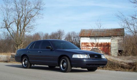 1998 Ford Crown Victoria - A dark blue sedan is parked on a dirt road with a rustic shed in the background under a clear sky.