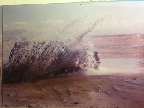 1988 Ford Bronco - A dark colored SUV is driving through a large splash of water, creating a dramatic spray. The vehicle is seen from the rear quarter angle,…