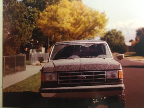 1988 Ford Bronco - A front 3/4 view of a dusty, dark-colored Ford Bronco with a cracked windshield parked on a street in front of trees with yellow foliage. T…