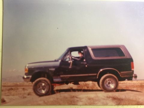 1988 Ford Bronco - A dark colored Ford Bronco is parked on a dirt embankment with a person visible in the driver's seat. The vehicle is viewed from the side w…