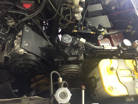 1988 Ford Bronco - Close-up view of a modified engine bay, showing an air conditioning compressor, pulleys, and a battery. A black metal bracket is mounted to…