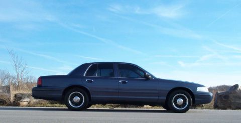 1998 Ford Crown Victoria - A dark blue sedan is parked on the side of the road under a bright blue sky with scattered clouds.