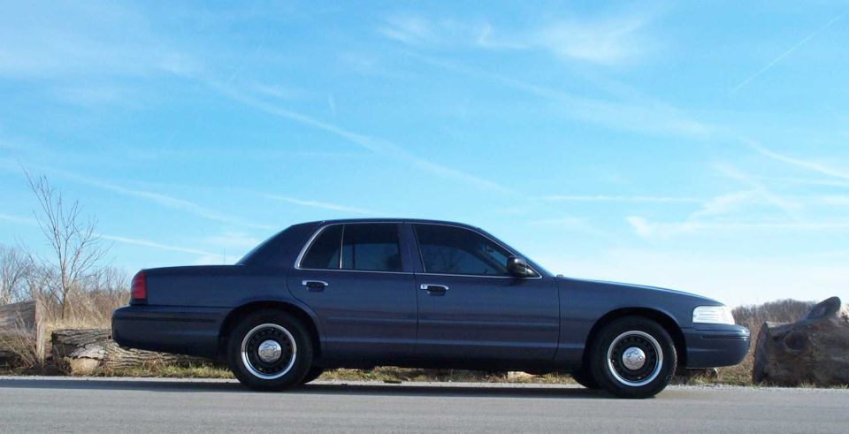 Side profile of a slate blue Ford Crown Victoria on black steel wheels parked beside large logs