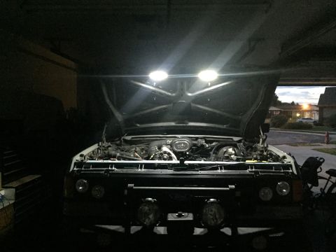 1988 Ford Bronco - Vehicle parked in a garage with hood raised and bright work lights illuminating the engine bay at dusk.