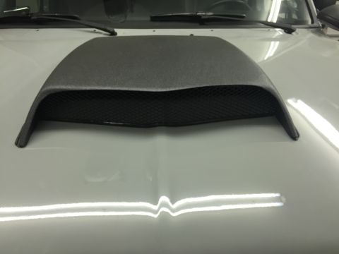1988 Ford Bronco - A close-up view of a black hood scoop with a mesh insert mounted on the hood of a white vehicle. The hood scoop has a textured finish and f…