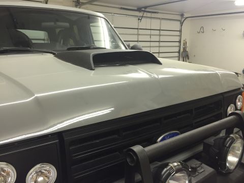 1988 Ford Bronco - A close-up, low-angle shot of the front of a white 1988 Ford Bronco with a black hood scoop and grille. The vehicle is parked in a garage w…