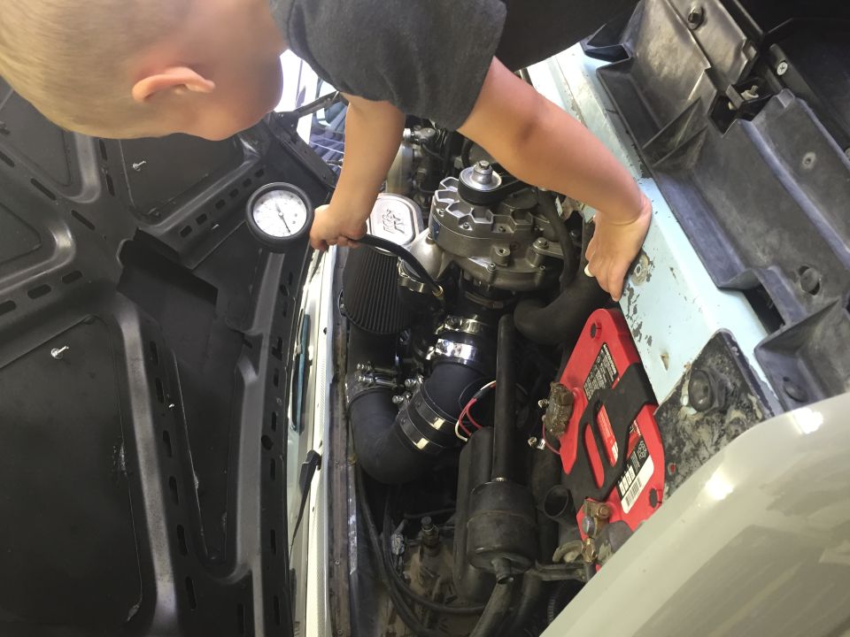 Child inspecting engine bay with pressure gauge. Visible: alternator, air intake, red battery.
