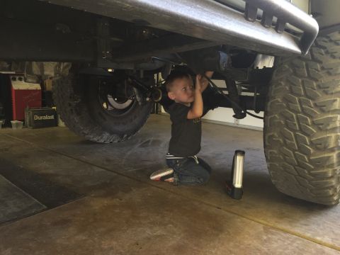 1988 Ford Bronco - A young boy kneels under the chassis of a large off-road vehicle, appearing to help with maintenance. A Duralast battery and a work light a…