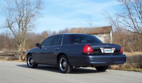 1998 Ford Crown Victoria - A dark blue sedan is parked on the side of a road with a rustic building in the background.
