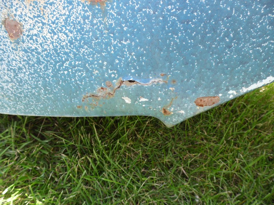 Close-up of faded blue body panel with rust holes and chipped paint above grass