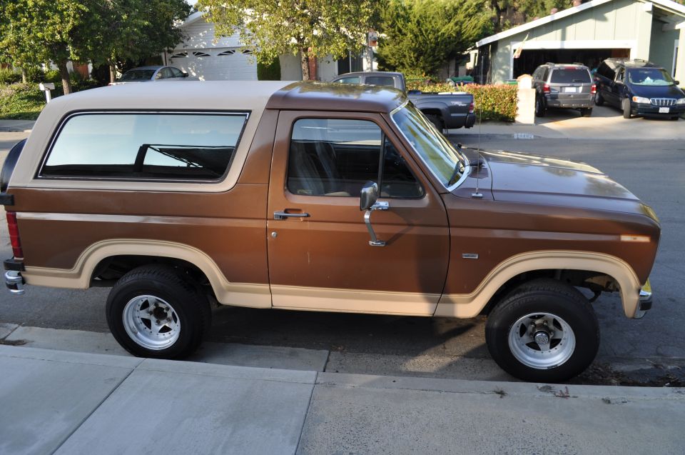Side view of a brown 1986 Ford Bronco with a tan top and aftermarket wheels parked on a street.