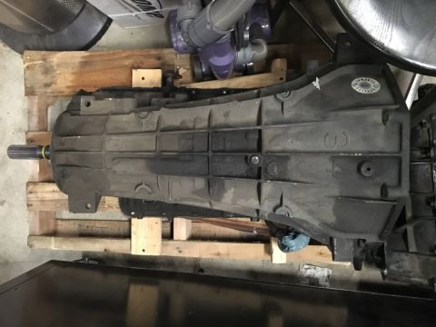 1993 Ford Bronco - A transmission assembly is shown from a bottom angle, resting on a wooden pallet. The transmission case is dark gray and shows some dirt an…