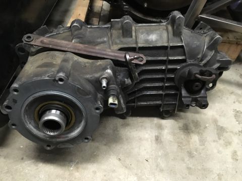 1993 Ford Bronco - A transfer case assembly is shown in a workshop setting. The cast aluminum housing has a ribbed texture and is bolted to a circular flange…