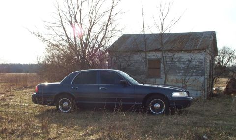 1998 Ford Crown Victoria - A 1998 Ford Crown Victoria sits idle in a rural setting, with an old shed and bare trees in the background.