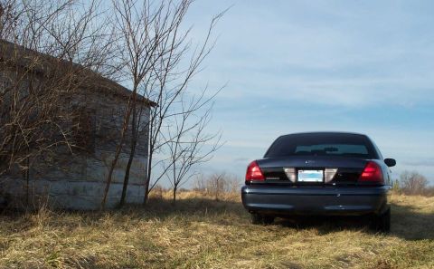 1998 Ford Crown Victoria - A dark 1998 Ford Crown Victoria is parked in a field next to a weathered, abandoned building under a partly cloudy sky.