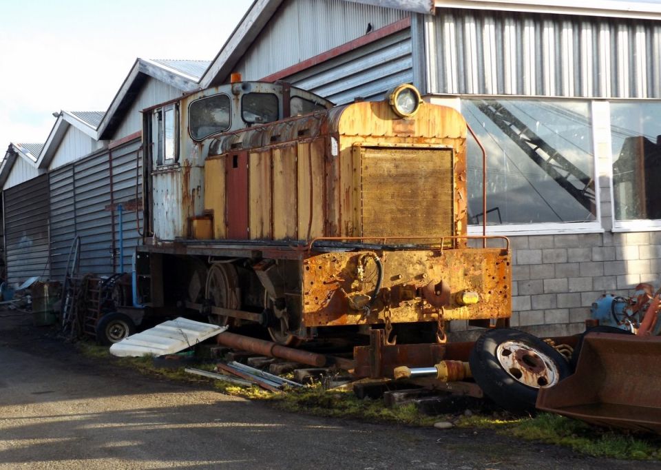 A rusty diesel locomotive parked outside a building with industrial parts around it.