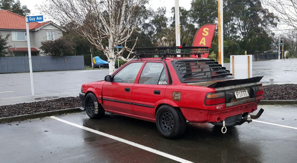 Modified red sedan with roof rack and spoiler parked in a wet lot.