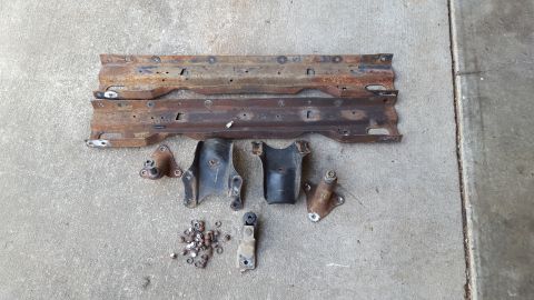 1994 Ford Bronco - Two rusty metal crossmembers are laid out on a concrete surface, along with several smaller brackets and a pile of fasteners. The crossmemb…