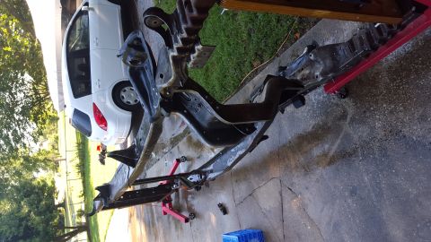 1994 Ford Bronco - A black vehicle frame is supported by red engine stands on a concrete driveway. A white wagon is visible in the background.