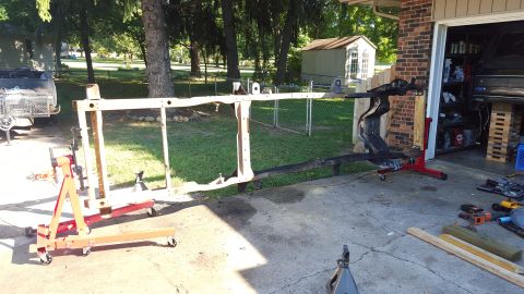 1994 Ford Bronco - A disassembled vehicle frame is supported by an engine hoist and jack stands on a concrete driveway in front of a garage. Tools and lumber…