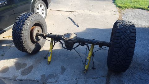 1994 Ford Bronco - A rear axle assembly with two large off-road tires and yellow shock absorbers is resting on a concrete driveway. The axle has a differentia…
