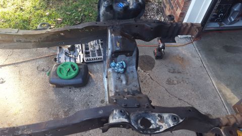 1994 Ford Bronco - A heavily rusted vehicle frame is laid out on a concrete driveway, surrounded by tools and automotive fluids. The frame appears to be under…