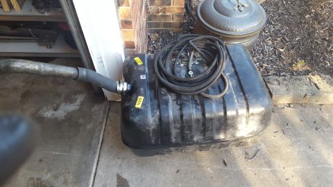 1994 Ford Bronco - A black plastic fuel tank with a coiled hose and fuel lines attached is resting on a concrete surface next to a brick wall. A metal pipe wi…