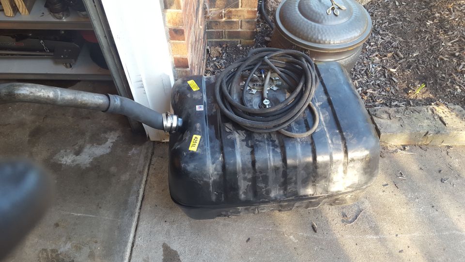 Black plastic fuel tank with coiled hose and fuel lines, attached to a metal pipe.