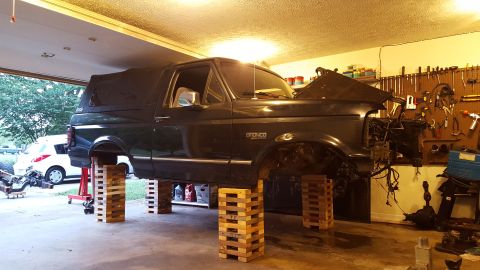 1994 Ford Bronco - A dark-colored Ford Bronco is elevated on wooden blocks in a garage. The front end of the vehicle is disassembled, with the hood open and e…