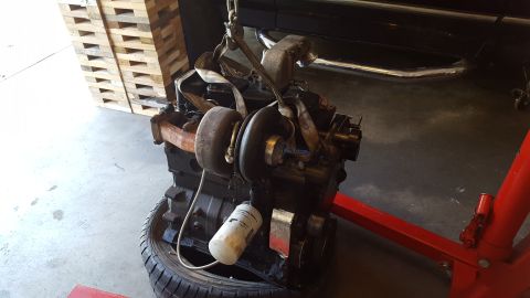 1994 Ford Bronco - A black engine with a turbocharger attached is suspended by straps and a chain, resting on a tire. A red engine hoist is visible supporting…