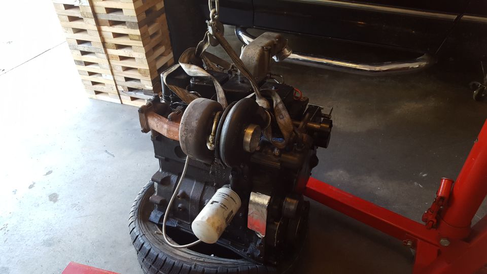 Black engine with turbocharger suspended by straps, resting on a tire, with a red engine hoist.