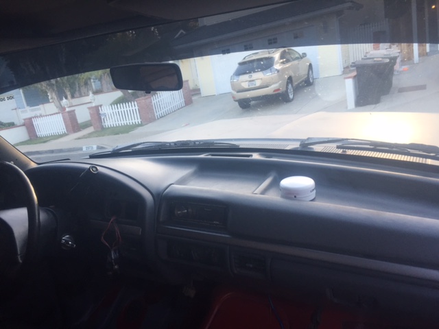 Interior view of a vehicle dashboard with a white device. Windshield shows a residential street with a gold SUV.
