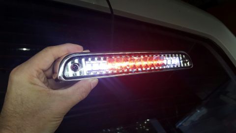 1996 Ford Bronco - A hand holds a chrome-trimmed LED third brake light assembly with red and white LEDs against the tinted rear window of a vehicle.