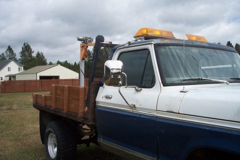 1973 Ford F-250 - A close-up side view of a blue and white flatbed truck with a wooden bed insert and a winch mounted on a boom. The truck has amber warning…