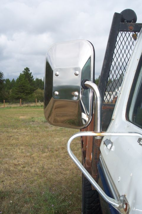 1973 Ford F-250 - A close-up view of a chrome side mirror mounted on the driver's side of a white truck. The mirror reflects a grassy field and trees under a…