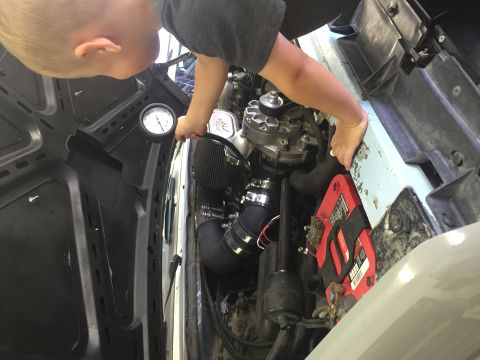 1988 Ford Bronco - A child leans over the engine bay of a vehicle, holding a gauge. Visible parts include a red battery, air intake, and part of the alternato…