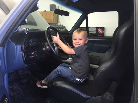 1988 Ford Bronco - A young boy sits in the driver's seat of a blue Ford Bronco, giving a thumbs up while holding the steering wheel. The interior shows black…