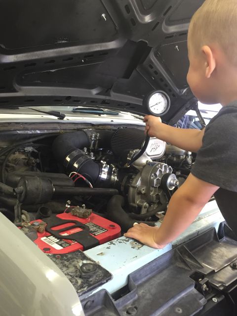 1988 Ford Bronco - A young child holds a pressure gauge over the engine bay of a vehicle, examining the supercharger and battery.