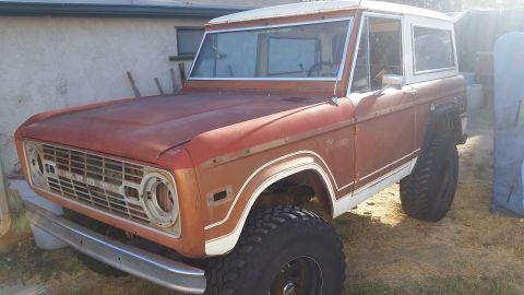 1973 Ford Bronco - A rusty, red 1973 Ford Bronco with a white hardtop is parked outdoors on a dirt and grass area. The vehicle features large off-road tires a…