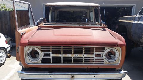 1973 Ford Bronco - The front view of a vintage, orange Ford Bronco with its hood and doors open, parked in front of a garage. The grille and headlights are vi…