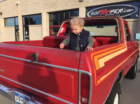 1978 Ford Bronco - Child sits in the bed of a red 1978 Ford Bronco, parked outside a building with a Softopper sign.