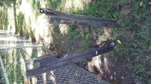 1994 Ford Bronco - Two dark metal car frame sections lie on the grass near a chain-link fence.