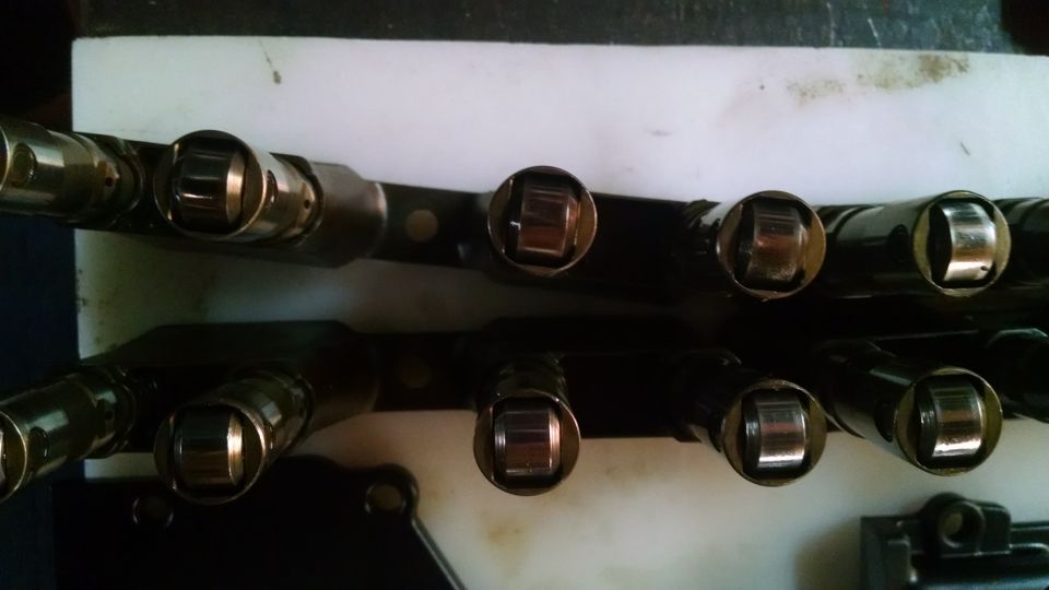 Two rows of metal rocker arms with roller tips, mounted on a black bar, against a white surface