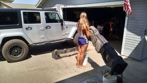 1985 Jeep CJ7 - A silver Jeep Wrangler is parked outside a garage while a woman holds a large black part.