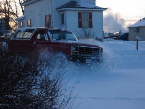 1981 Ford Bronco - Red SUV driving through deep snow in a residential area during winter.
