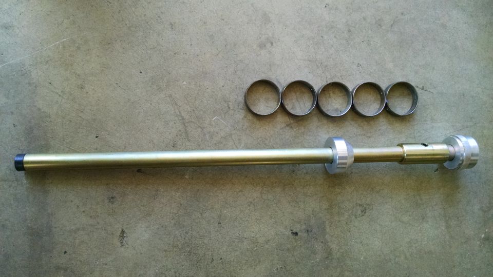 Metal tool with a long shaft, adjustable rings, and a cap, next to five loose rings on a concrete surface