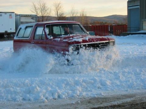 1981 Ford Bronco - A red SUV navigates a snow-covered area, creating a large spray of snow.