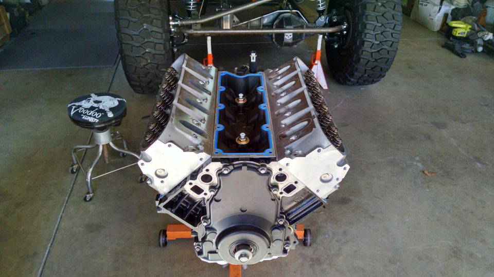 V8 engine on an orange stand, with a Jeep's rear tire and suspension visible behind it. A stool with 'Voodoo' text is to the left