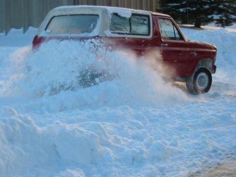 1981 Ford Bronco - A red SUV drives through deep snow, spraying it up around the tires.